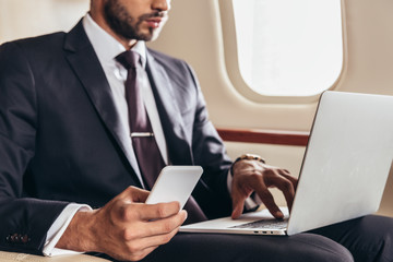 cropped view of businessman in suit using laptop and smartphone in private plane