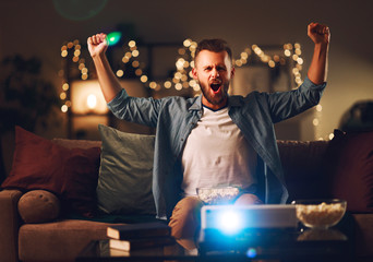 young male cheerleader watching football on tv projector at home in evening