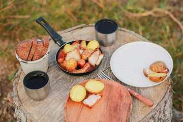 Roasted potatoes in rind with bacon, meat. Fried potatoes in a frying pan in the woods. Bread fried on a fire. Camping with food. Picnic on a stump with tea. Fried sausages. To eat in nature