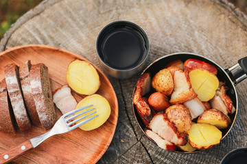 Roasted potatoes in rind with bacon, meat. Fried potatoes in a frying pan in the woods. Bread fried on a fire. Camping with food. Picnic on a stump with tea. Fried sausages. To eat in nature