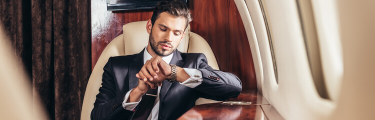 panoramic shot of handsome businessman in suit looking at wristwatch in private plane