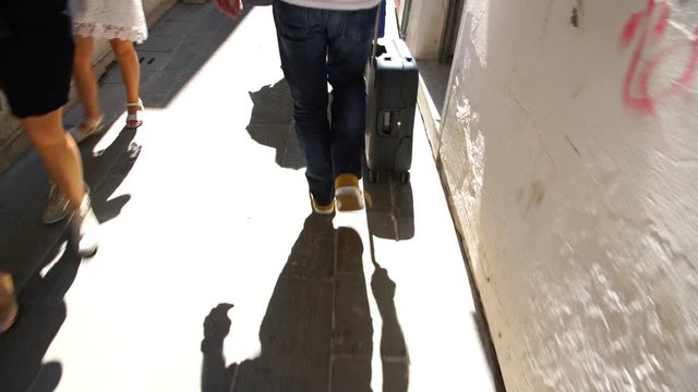 Man In Blue Jeans And White Shirt Pushes Suitcase Down Narrow Street Passing Through Crowd In Venice Backside View