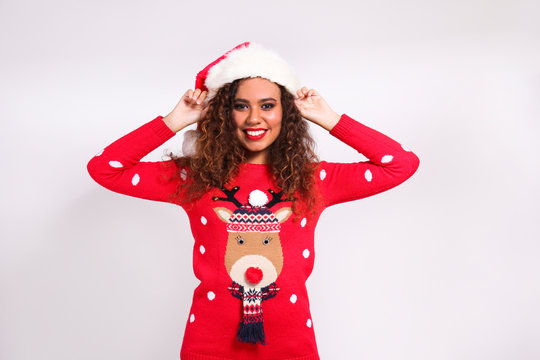 Studio portrait of young woman with dark skin and long curly hair wearing tight santa claus hat and christmas outfit. Ugly sweater concept. Close up, copy space for text, isolated background.