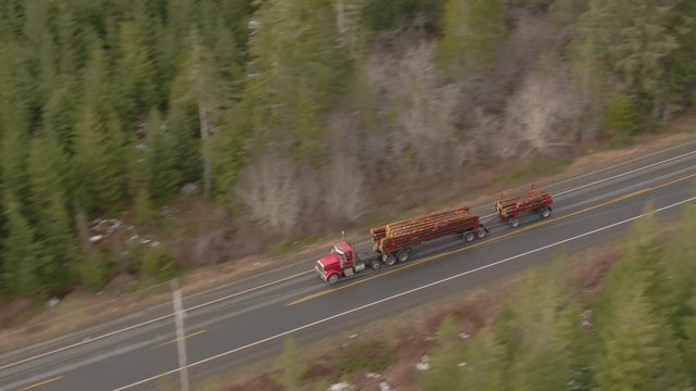 DRONE: Flying above two large trucks as they transport logs down the scenic rural highway crossing the picturesque Olympic Natural Park. Big rigs transport logs down freeway leading through a forest.