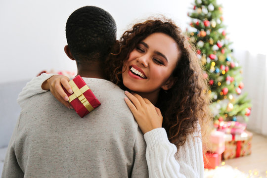 Portrait of young couple, boyfriend & girlfriend with dark skin wearing santa claus hat and christmas outfit exchanging gifts at home. Close up, copy space for text, isolated background.