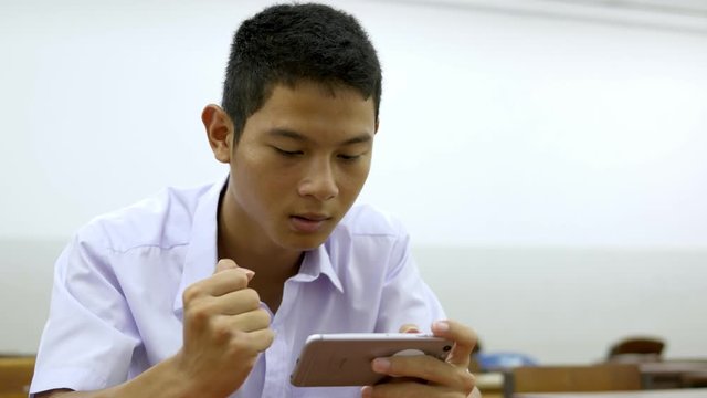 Asian male high school student in a white uniform, who is a lot of addicted to games. He is playing exciting games on  their mobile phones and sitting in the classroom.