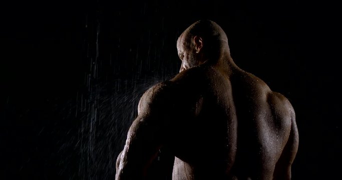 Back brutal muscular male bodybuilder close-up on a black background, he is in the rain, water flows down it.
