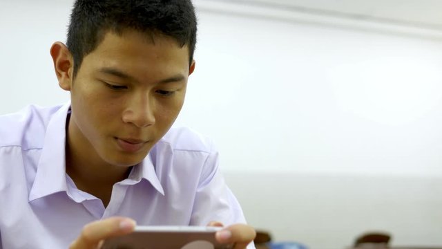 Asian male high school student in a white uniform, who is a lot of addicted to games. He is playing exciting games on  their mobile phones and sitting in the classroom.