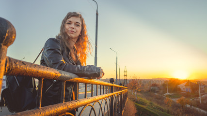A young beautiful woman in a leather jacket with a black backpack stands thoughtfully on the bridge against the background of the urban district.