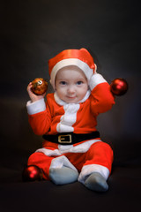 Fototapeta na wymiar Little baby dressed as Santa Claus sits on a dark background and plays with Christmas toys