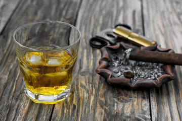 A glass of whisky on old wooden table. Near in defocusing lies a smoldering cigar on an ashtray and guillotine with lighter.