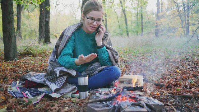 Woman camping in a forest, sitting at a campfire. in sunset at autumn. Young caucasian woman speaking on phone.