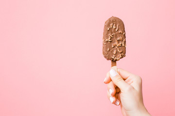 Young woman hand holding ice cream with nuts and chocolate glaze on pastel pink background. Empty...