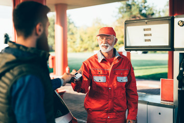 Young handsome adult man together with senior worker standing on gas station and fueling car.