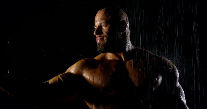 Portrait of a brutal muscular bald bearded male bodybuilder close-up on a black background, he poses, shows biceps, he is in the rain, water flows down him.