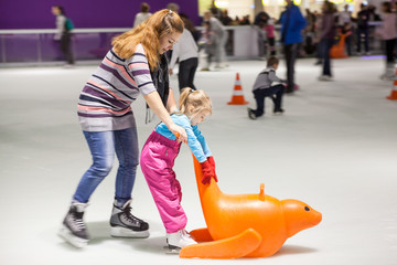 Mother dressed sweater, jeans and scarf helping her daughter to ice-skating, indoor ice-scating...