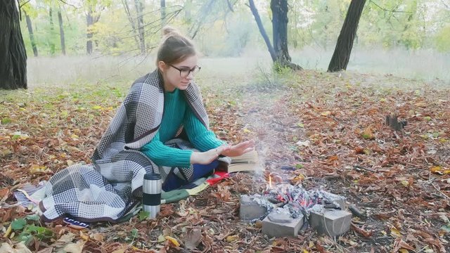 The yong caucasian woman in a warm green sweater sitting by the fire in the evening in foreat at autumn.