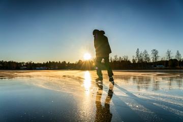 Boy silhouette against of the Sun: a teenager is ice skating on the crystal clear frozen lake - ice surface like big mirror. Low sun lights with warm light at very cold winter day, Northern Sweden - obrazy, fototapety, plakaty