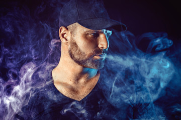 Brutal bearded man vaping and releases a cloud of vapor. Dark colored background.