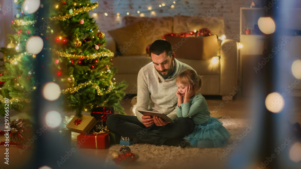 Wall mural looking through the window. father reads book to his daughter, sitting under christmas three with gi