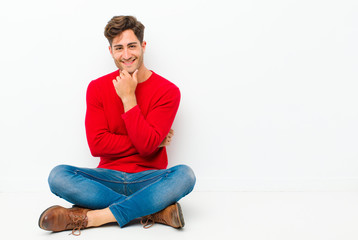 young handsome man looking happy and smiling with hand on chin, wondering or asking a question,...