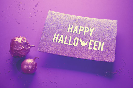 Creative pink and purple Halloween background on neon paper with glittering pumpkin painted metallic. Spotlight shining from underneath glittering paper card with text "Happy Halloween".