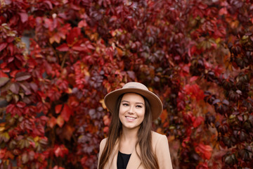 Fototapeta na wymiar Young woman with a sincere smile having a good time an autumn day. Closeup portrait of charming lady in beige classic coat and hat on background of bright autumn leaves. 