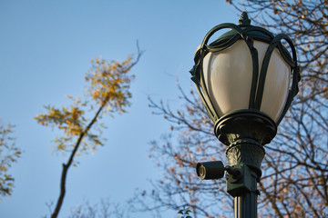 Vintage lantern with a surveillance camera in a public park in autumn in front of a blue sky, selective focus