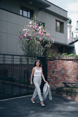 Blonde confident female in classical grey suit going from her modern house. Young girl looking away and holding jacket in hand. Concept of fashion and business.