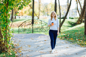 Young attractive woman enjoy a cup of morning coffee in autumn park. Slim blonde girl with beautiful hair in white sweater drink takeaway latte at forest