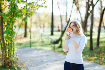 Young attractive woman enjoy a cup of morning coffee in autumn park. Slim blonde girl with beautiful hair in white sweater drink takeaway latte at forest