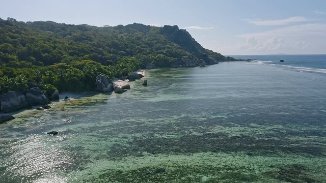 Aerial footage of tropical paradise beach Anse Source d'Argent. Shallow lagoon with beautiful algae pattern, granite rocks and rainforest around in morning sun light. La Digue Island, Seychelles
