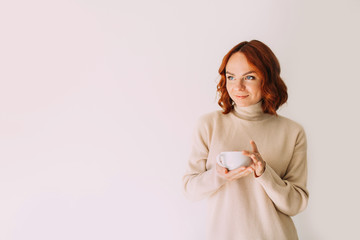 Lifestyle portrait of red haired beautiful woman drinking coffee from a white mug, wearing cozy sweater.