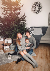 Christmas family with baby smiling near the Xmas tree. Living room decorated by Christmas tree and present gift boxes, the light give cozy atmosphere. New Year holidays