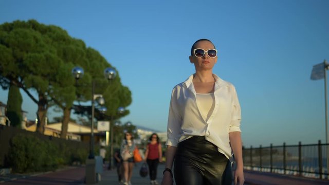 stylish bald girl in sunglasses and a white shirt walks the street against a blue sky and green trees
