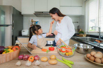 Portrait of beautiful Asian young woman and her daughter cooking salad for lunch and feeding tomato to her mother while making food, Family life love relationship, or home fun leisure activity concept