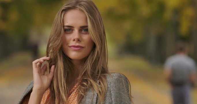 Close-up portrait of beautiful Caucasian woman standing in autumn park and tucking hair. Pretty girl in checkered jacket and mustard dress turning to camera and smiling. Cinema 4k footage ProRes HQ.