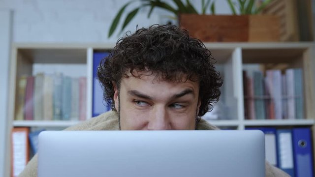 Funny caucasian man with curly hair hiding head behind monitor spying his colleagues working by computer in corporate office. Close-up.