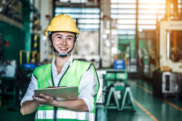 Asian woman is industrial engineer or qc team holding th clipboard while standing in the heavy duty manufacturing shop floor. Factory worker concept.