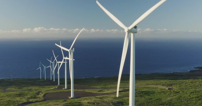 Aerial static shot of a row of windmills spinning at sunrise with green fields and ocean in the background, wind energy farm generating electricity, green energy future