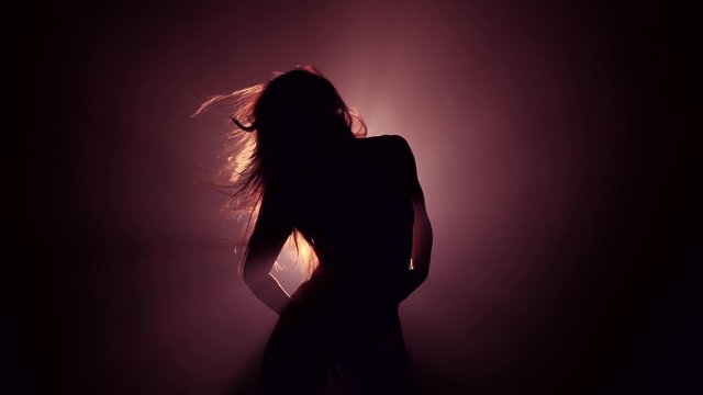 Silhouette of beautiful dancer on smoky dark background.Spotlight shines back against sexy girl. She moving seductively.