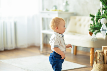 Christmas. A small blond boy in a shirt and blue jeans stands next to a Christmas tree with toys in a bright room.