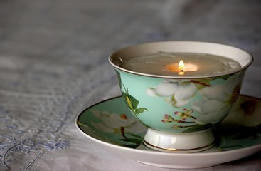 A china cup filled with a lit soy candle is placed on a table covered with an antique linen with blue embroidery