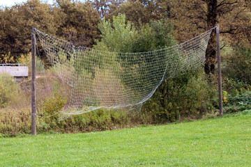 Broken large white protection net tied to two strong wooden poles behind local playground surrounded with freshly cut grass and dense trees in background on warm sunny autumn day