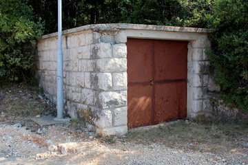 Fototapeta na wymiar Detached small garage with rusted metal doors and stone walls next to gravel road surrounded with dense trees and vegetation on warm sunny summer day