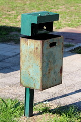 Dilapidated partially rusted green metal trash can with faded color mounted on metal pole with concrete foundation next to stone tiles path at local public park on warm sunny summer day
