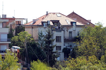 Old family house villa in need for restoration with dilapidated facade and new roof tiles surrounded with dense trees and other family houses on warm sunny summer day