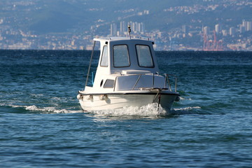 Tall small white fishing boat with open cabin coming in local harbour surrounded with blue sea and dense city in background on warm sunny summer day