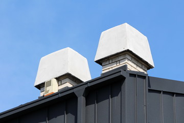 Two white brick chimneys with large concrete finish on top of modern apartment building metal roof with alarm notification device on one side and clear blue sky in background