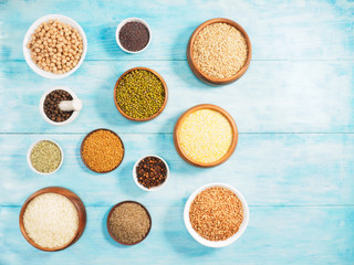 Rice, green gram, allspice, chick pea, allspice, buckwheat, oat, cloves, fenugreek, fennel, mustard seeds in cups and spoons on blue wooden background